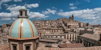 Сицилия без хайпа Case a 1 euro в Кальтаджироне: официальный перезапуск к середине 2026 Кальтаджироне перезапуск проекта «дом за 1 евро» к середине 2026 Исторический центр Кальтаджироне (Сицилия), фасады и лестницы с керамикой, на переднем плане папка «Case a 1 euro candidatura»